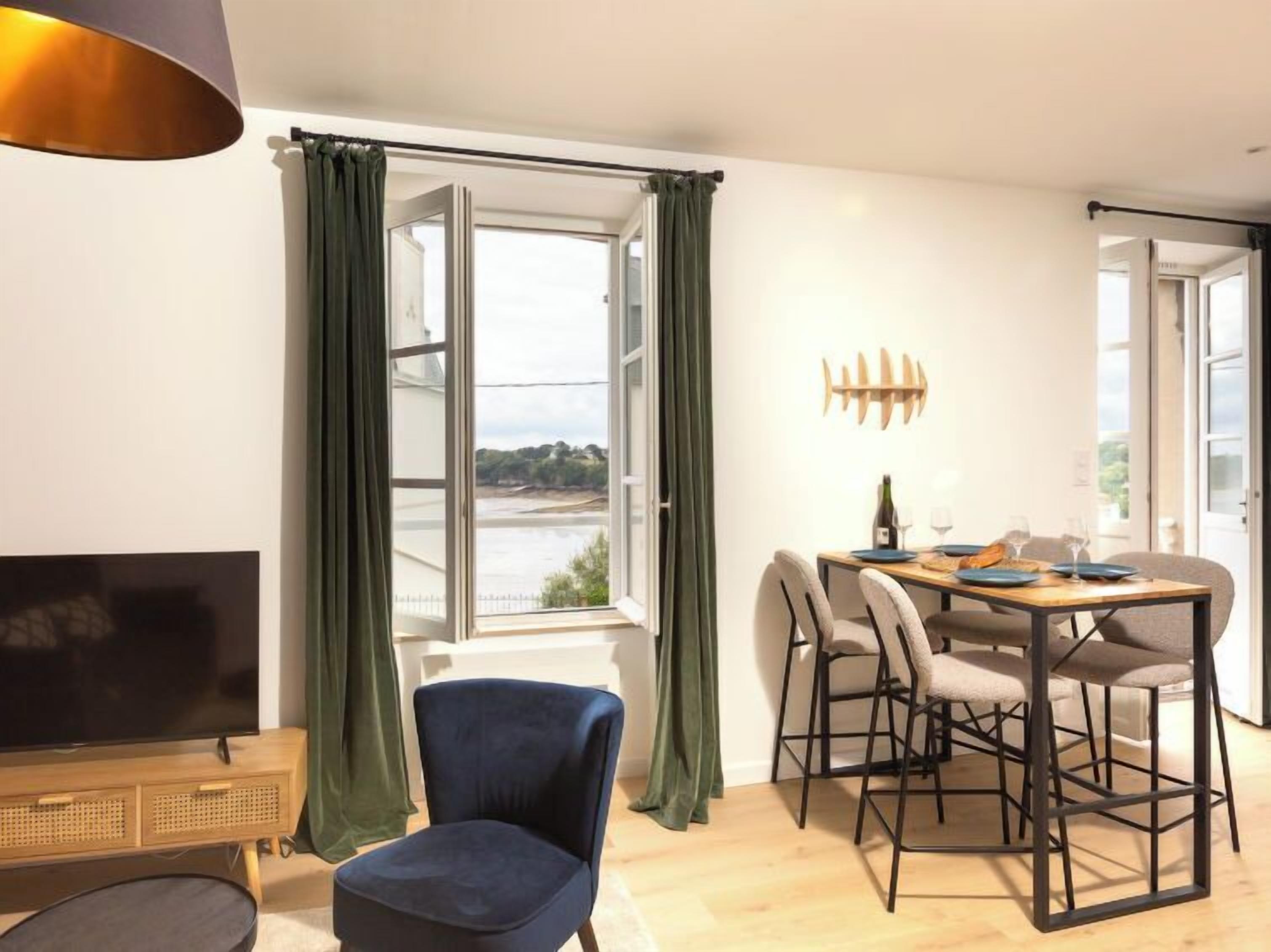 A modern living room with a television, a dining table with chairs, a large window with curtains, and a view of the sea.