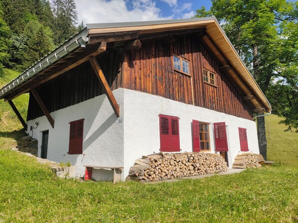 Une maison rustique en bois, à l’extérieur peint en blanc et dotée de volets rouges, située sur une colline verdoyante.