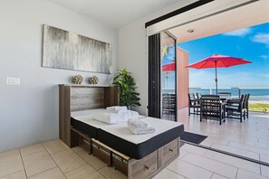 A modern bedroom with a large bed, a potted plant, and a view of the beach through an open door.