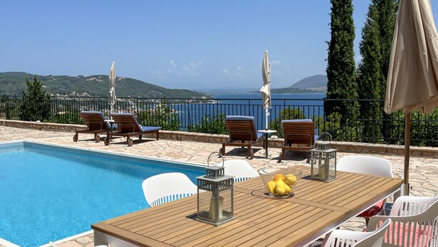 A poolside area with a wooden table, white chairs, and a view of the sea. - Villa Fortuna in Nikiana (Lefkada)