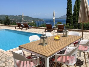 Ein Poolbereich mit Holztisch, weißen Stühlen und Blick auf das Meer.