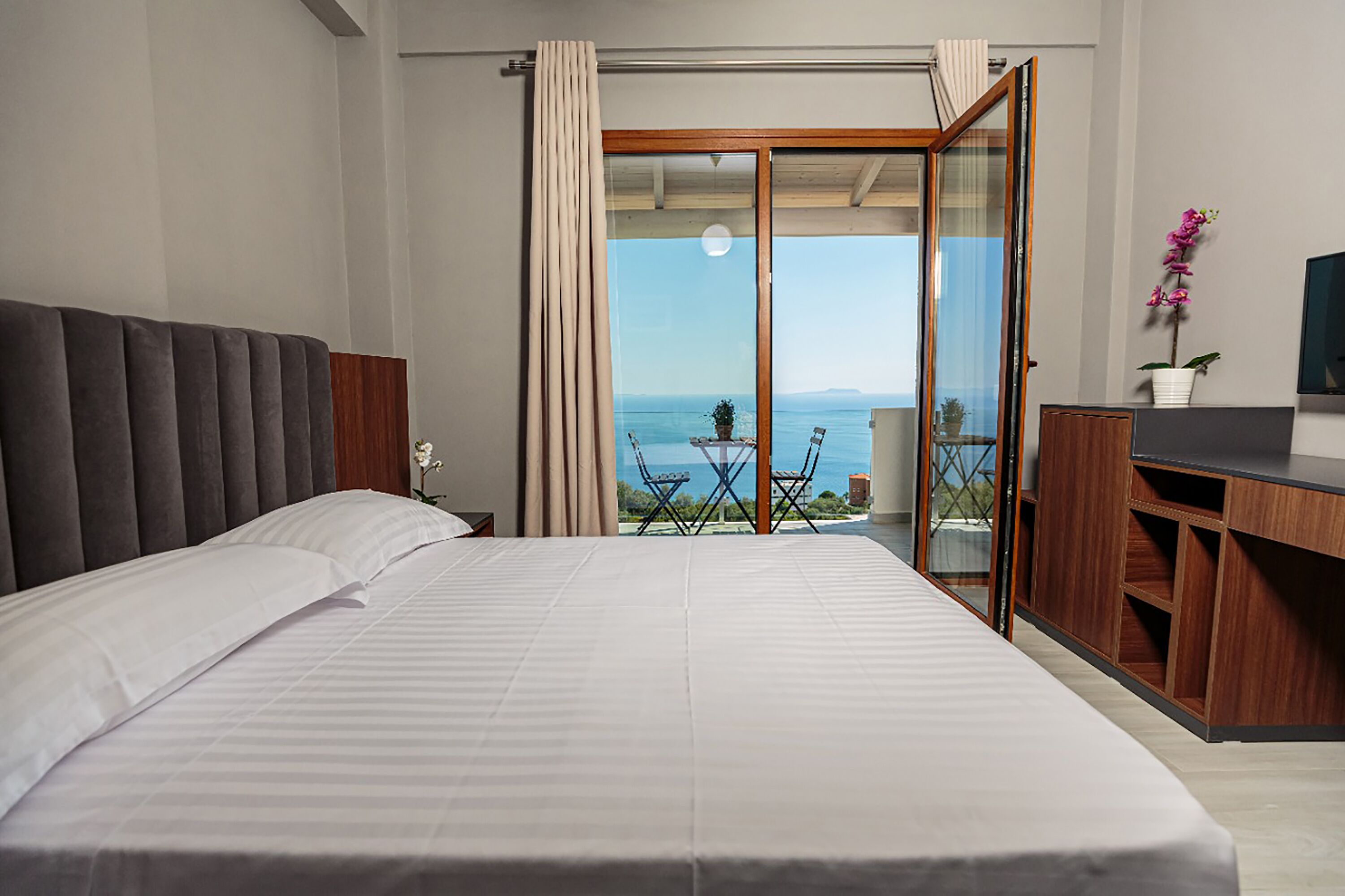 A bedroom with a large bed, a wooden desk, a television, and a balcony with a view of the sea.