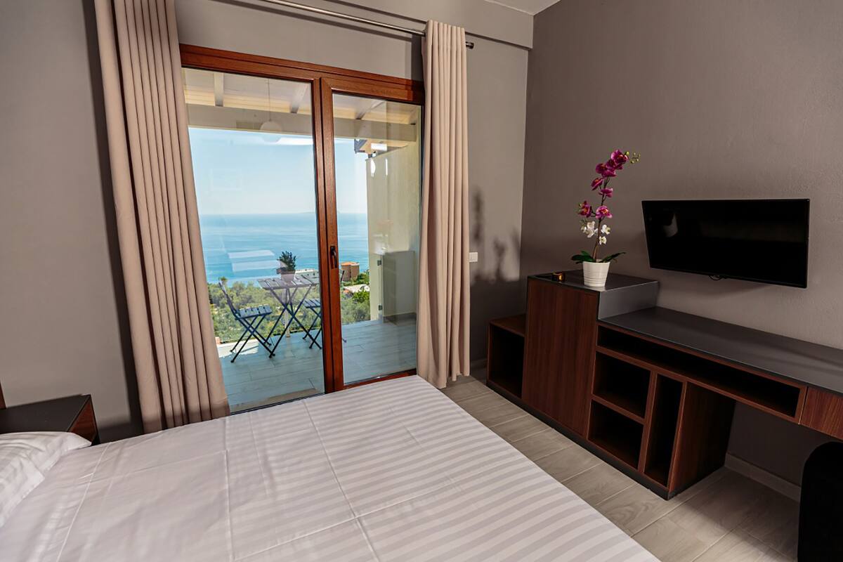 A hotel room with a bed, a TV, a desk, and a balcony with a view of the ocean.