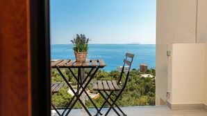 Un balcón con una mesa y sillas de madera, una planta de mimbre y vistas al mar y edificios lejanos.