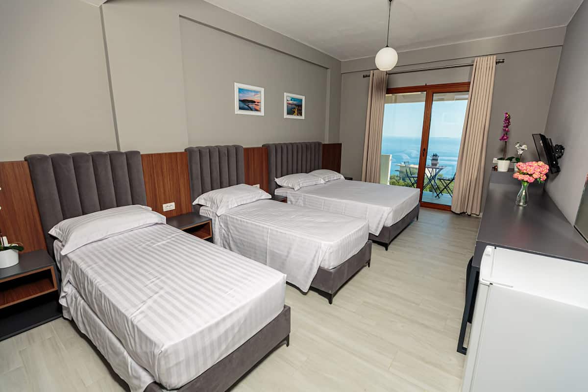 A hotel room with three beds, a small kitchen area, and a balcony with a view of the sea.