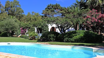 A swimming pool surrounded by lush greenery and a white building with an arched entrance.