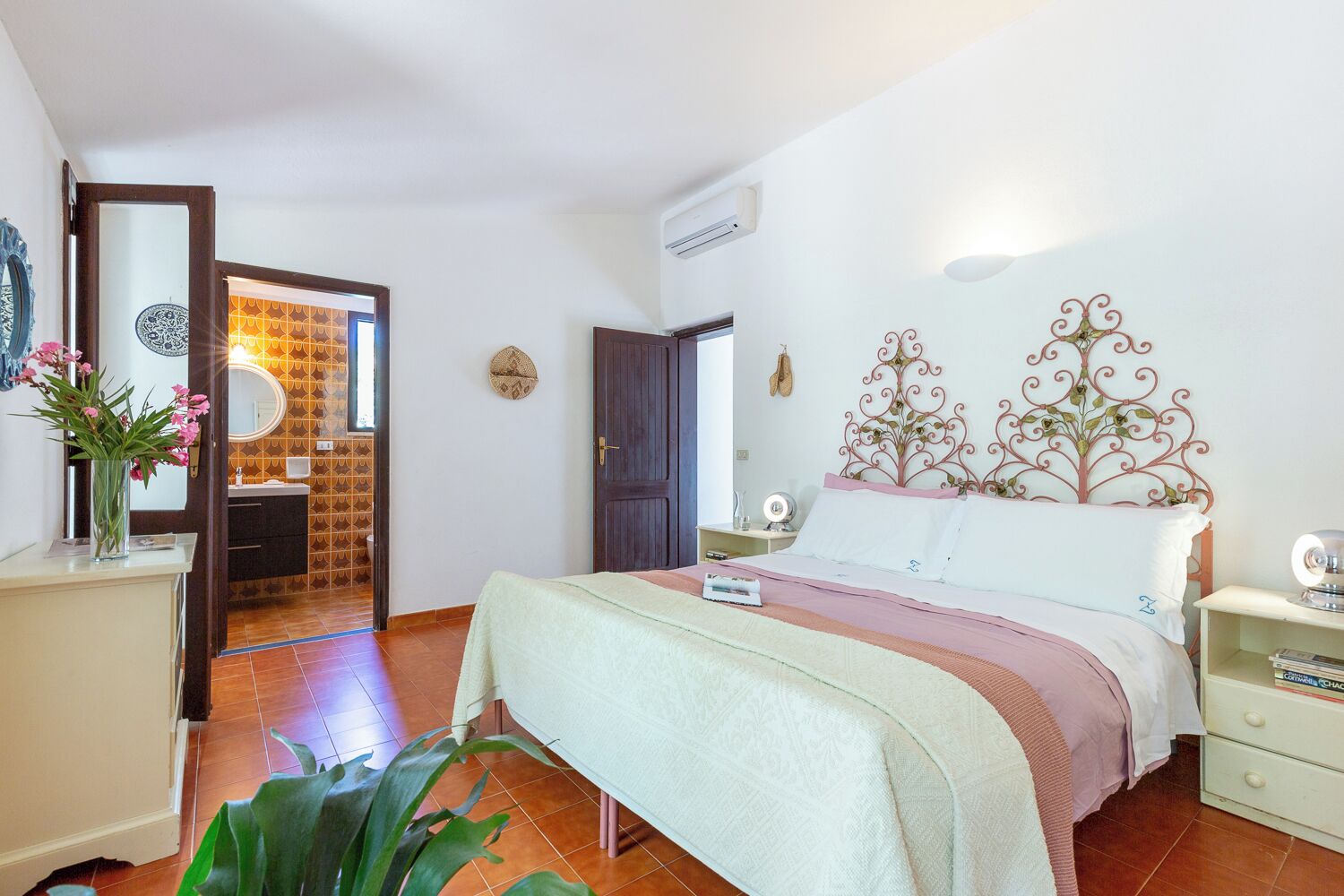 Quarto com cama, mesa de cabeceira e criado-mudo. Banheiro visível por uma porta aberta.