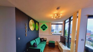 A modern living room with a green sofa, a black ottoman, and a large window offering a city view. - Bull's Ring 3bed Penthouse City Centre (Birmingham)