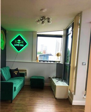 Un salon moderne avec un canapé vert, un petit pouf vert, une fenêtre donnant sur des immeubles et un néon mural.