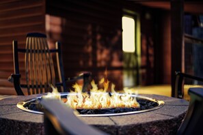 Feuerstelle mit Flammen, umgeben von Stühlen, in einer Außenanlage mit einer Holzverkleidung im Hintergrund.