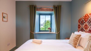 Ein Schlafzimmer mit Bett, einem Fenster mit Vorhängen, Blick auf Grünfläche und Wandbehang mit Muster.