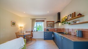 A compact kitchen with a dining area, featuring a window with curtains, a built-in stove, and wooden shelves above the counter.