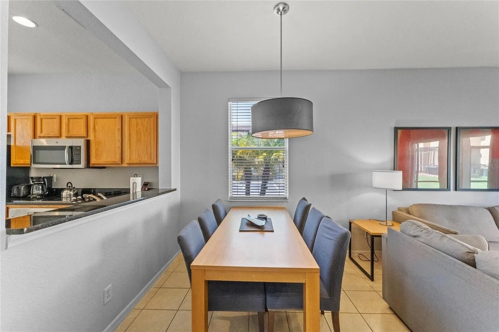 A modern kitchen with wooden cabinets, a microwave, and a coffee maker. There is a dining table with chairs and a grey sofa. A pendant light hangs above the table.
