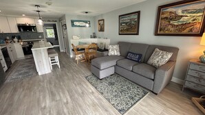 A modern living room with a grey sofa, a dining table with chairs, a kitchen area with white cabinets, and a large painting on the wall. - Ocean Towers 111 Condo (Ogunquit)