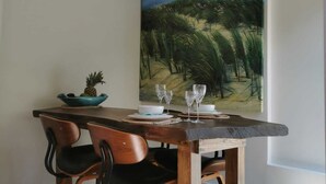 A dining area with a wooden table, two chairs, a painting of a landscape, and a ceiling fan.