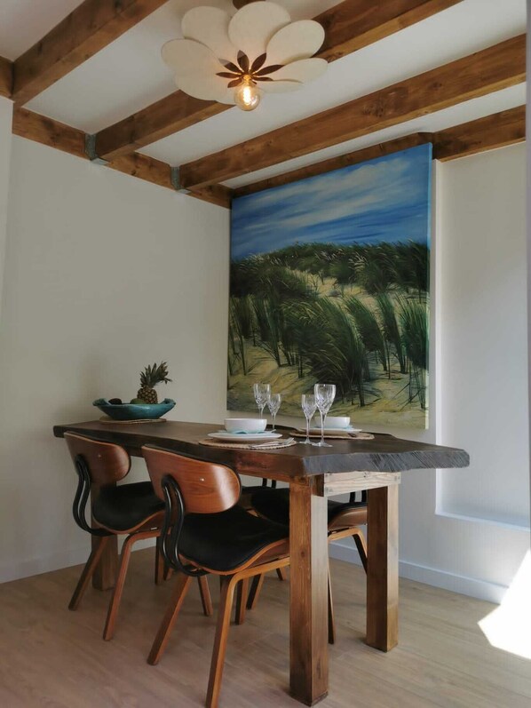 A dining area with a wooden table, two chairs, a painting of a landscape, and a ceiling fan.