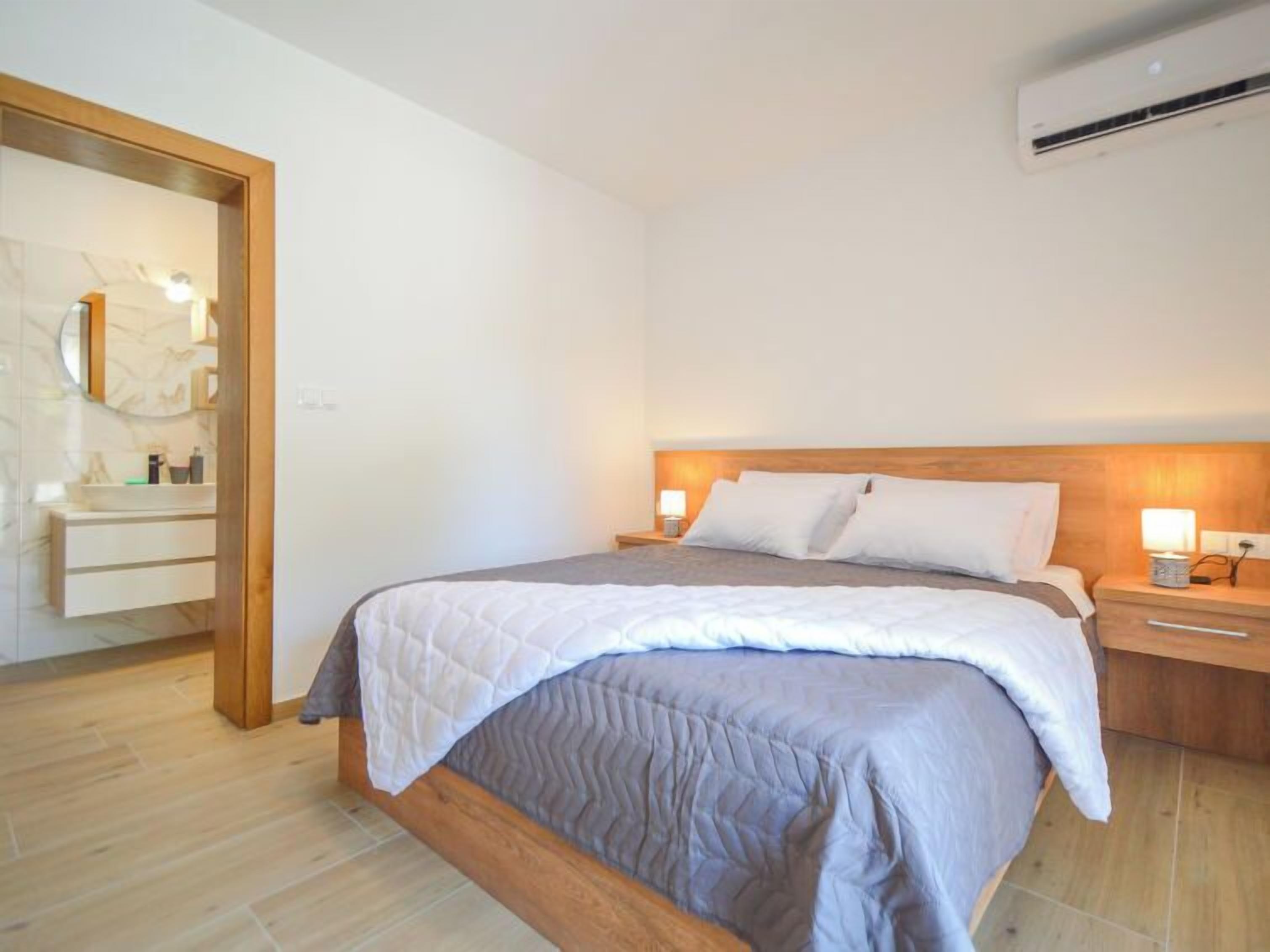 Une chambre à coucher moderne avec une tête de lit en bois, un grand lit recouvert d’une couette grise, des tables de chevet avec des lampes, et une salle de bain visible à travers une porte ouverte.