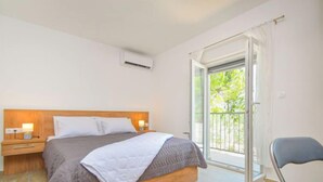 Een slaapkamer met een bed, nachtkastjes, een airconditioning, een stoel en een balkon met uitzicht op groen.
