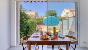 Un coin repas avec une table dressée pour deux personnes, un parasol bleu et une vue sur le quartier, visible à travers de grandes portes-fenêtres coulissantes.
