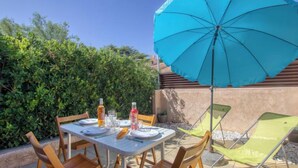 Un espace extérieur aménagé pour les repas, avec une table dressée pour quatre personnes, entourée de chaises en bois. Un grand parasol bleu offre de l’ombre.