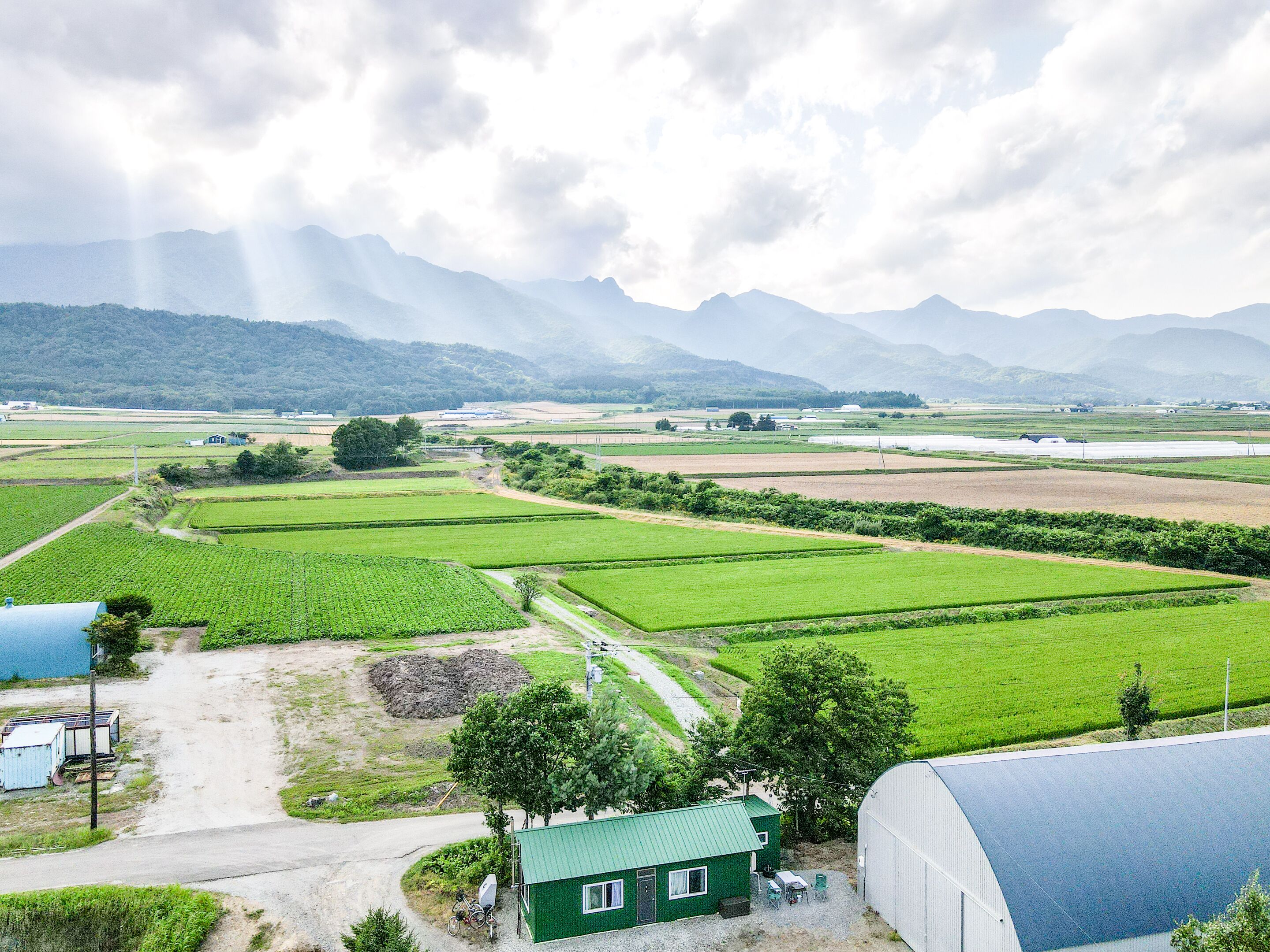 A rural landscape with fields, a small house, and mountains in the background.