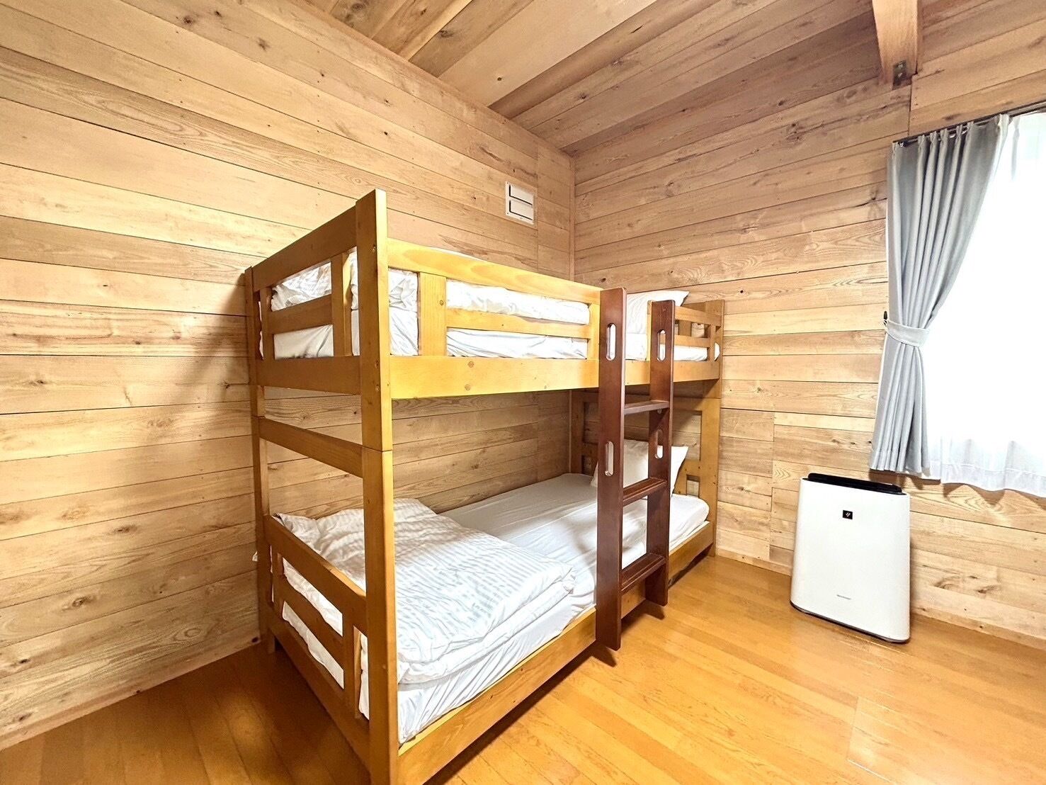 A wooden bunk bed room with a small white appliance, wooden walls, and a window with curtains.