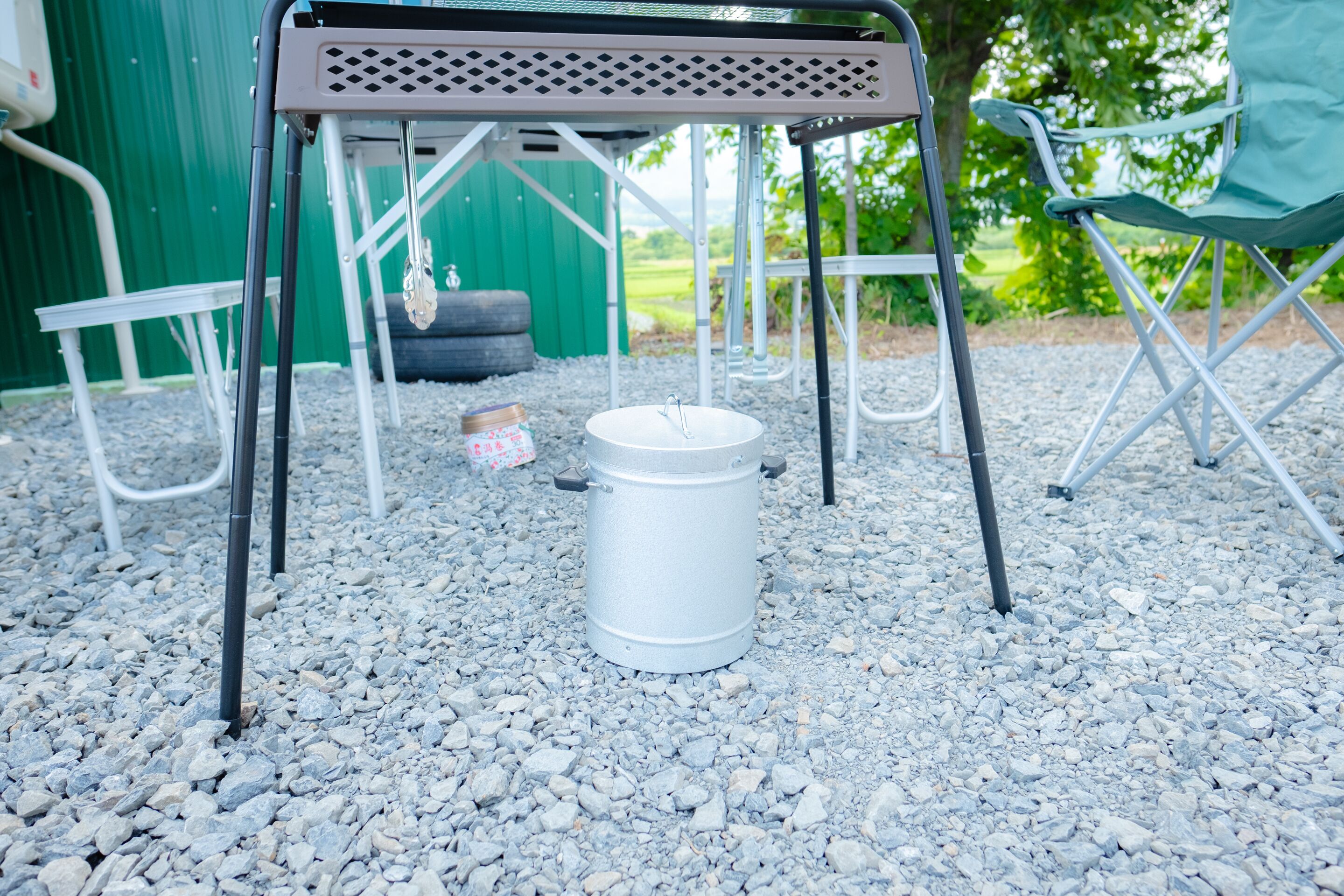 A metal table with a metal frame, a white bucket, and a green chair in a gravel area.