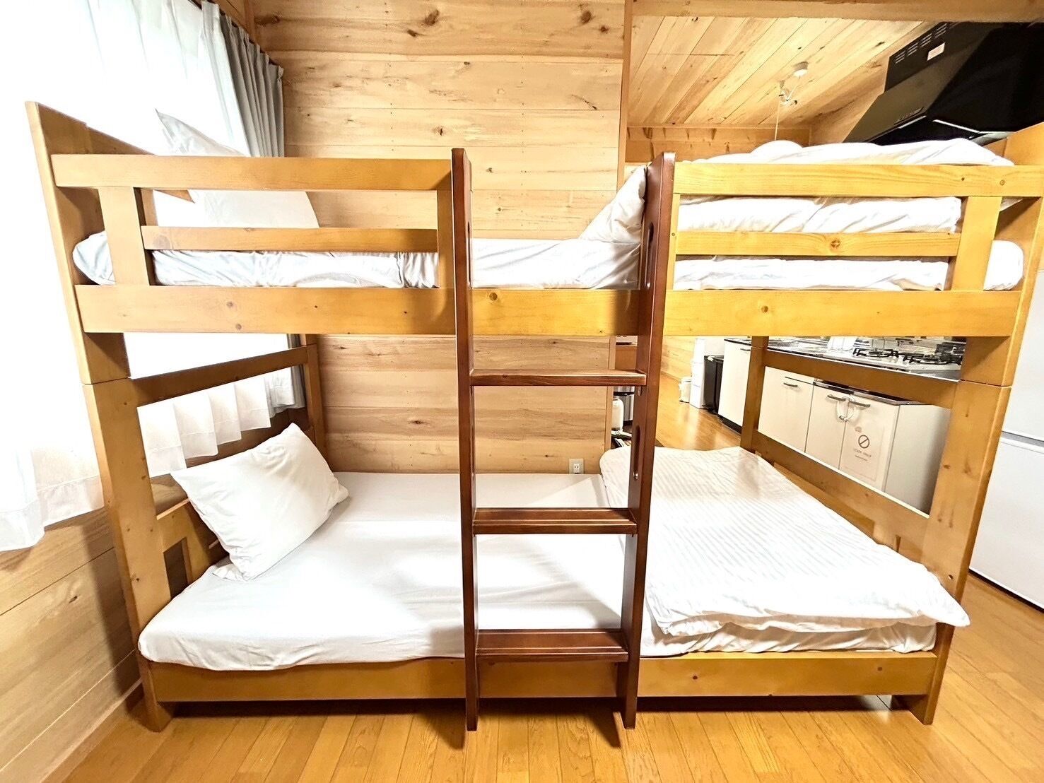 A bunk bed room with a wooden ladder, a small kitchen area, and a window with curtains.