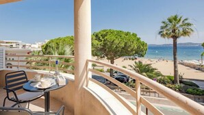 A balcony with a table and chairs, overlooking a beach with palm trees and a clear sky. - Les Dauphins by Interhome (Sainte Maxime)