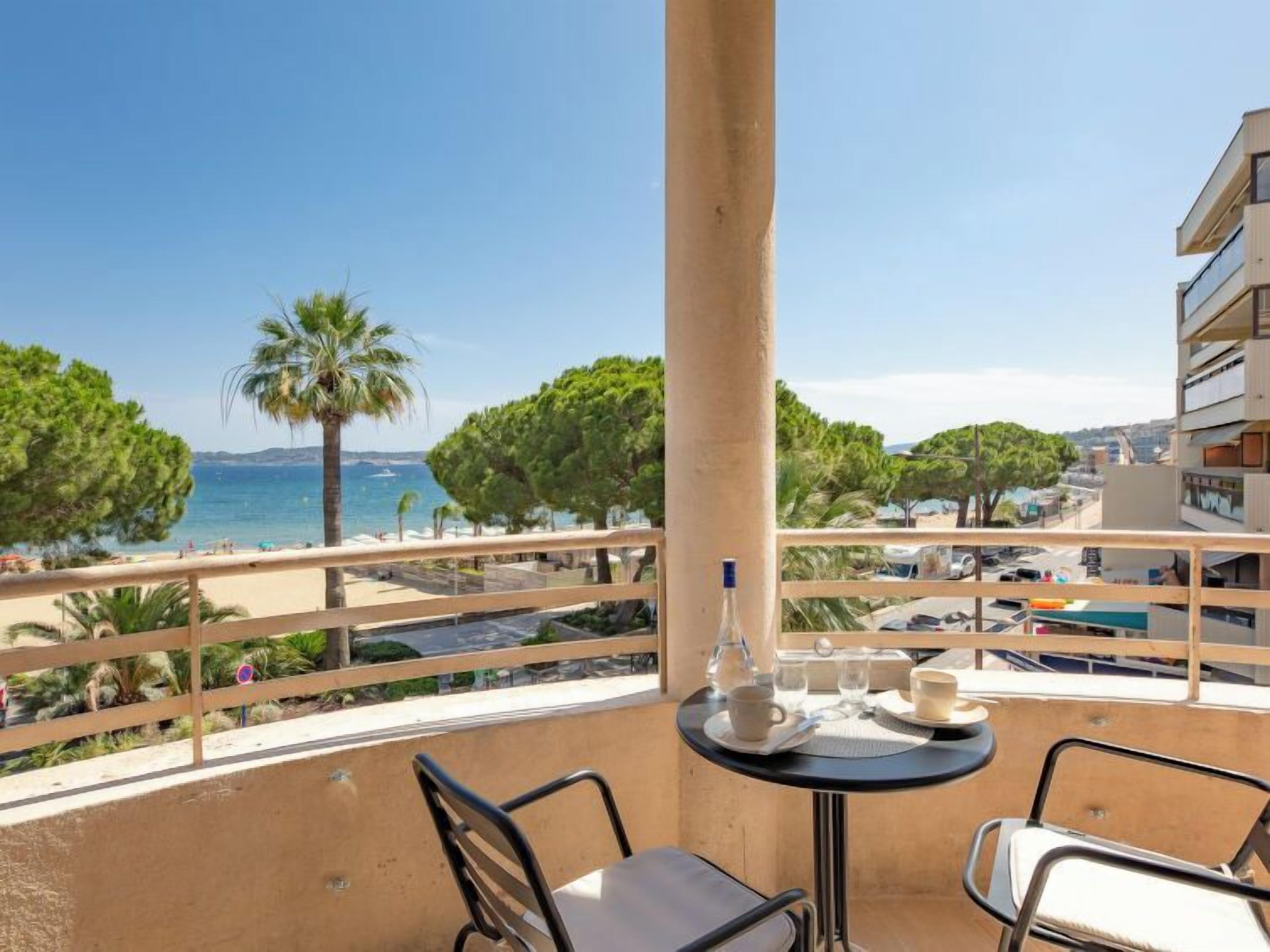 Ein Balkon mit Tisch für zwei Personen mit Blick auf einen Strand, an dem Palmen stehen, und einem strahlend blauen Himmel.