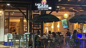 Un área de estar al aire libre en el restaurante LaKaza, con mesas, sillas, sombrillas y una barra visible al fondo.