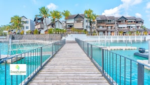 Un muelle de madera que lleva a una zona de piscina con varios edificios al fondo.