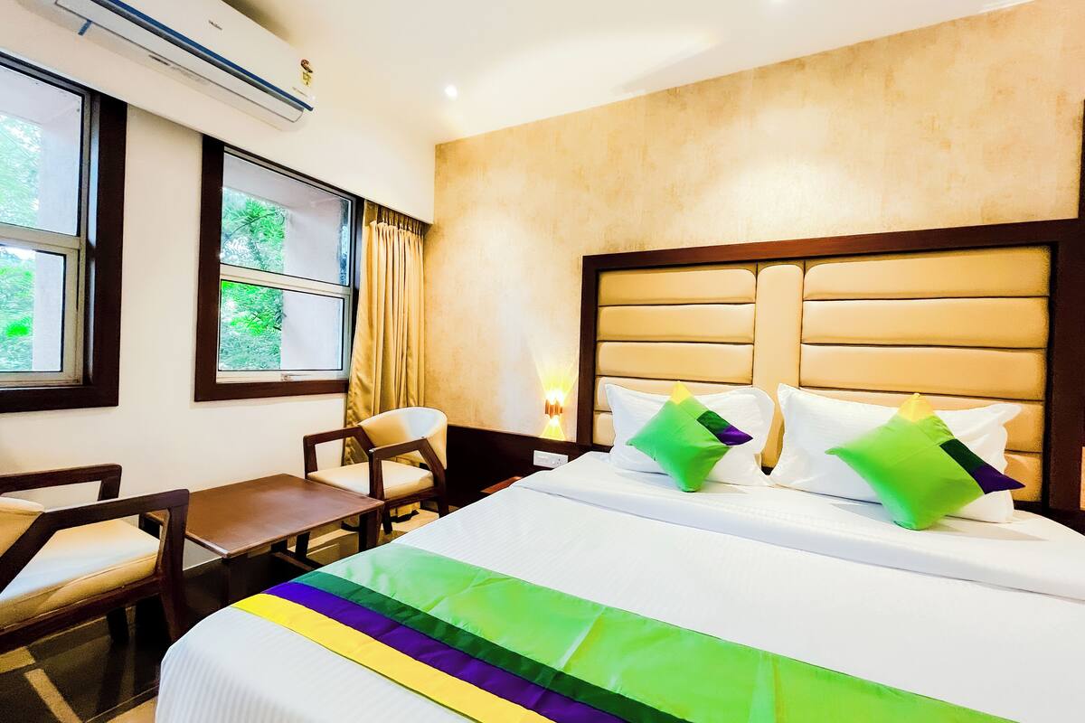 A hotel room with a bed, a desk, and a chair. There is a window with curtains, an air conditioning unit, and a wall-mounted lamp.