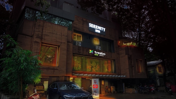 Un edificio con un letrero que dice 'Serenity Inn' y el logo de 'Treebo', con un estilo arquitectónico moderno, amplias ventanas y un área de estacionamiento al frente.
