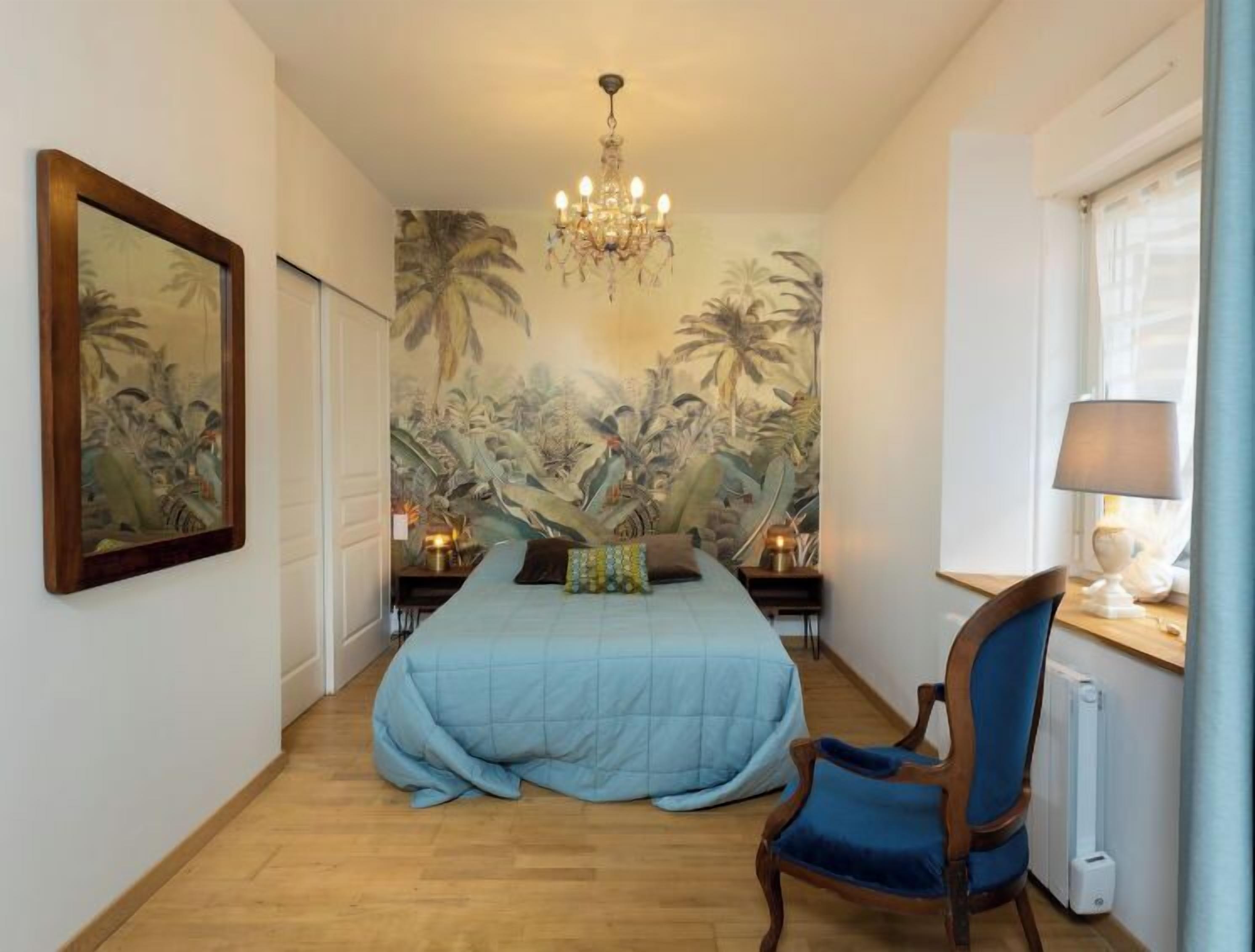 A bedroom with a bed, a chair, a chandelier, a wall mural, and a window with a lamp.