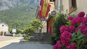 A building with a sign reading "CAFETERIA" and flags, a paved area with parked cars, and mountains in the background. - Restaurante Hotel BADAIN (Tella)