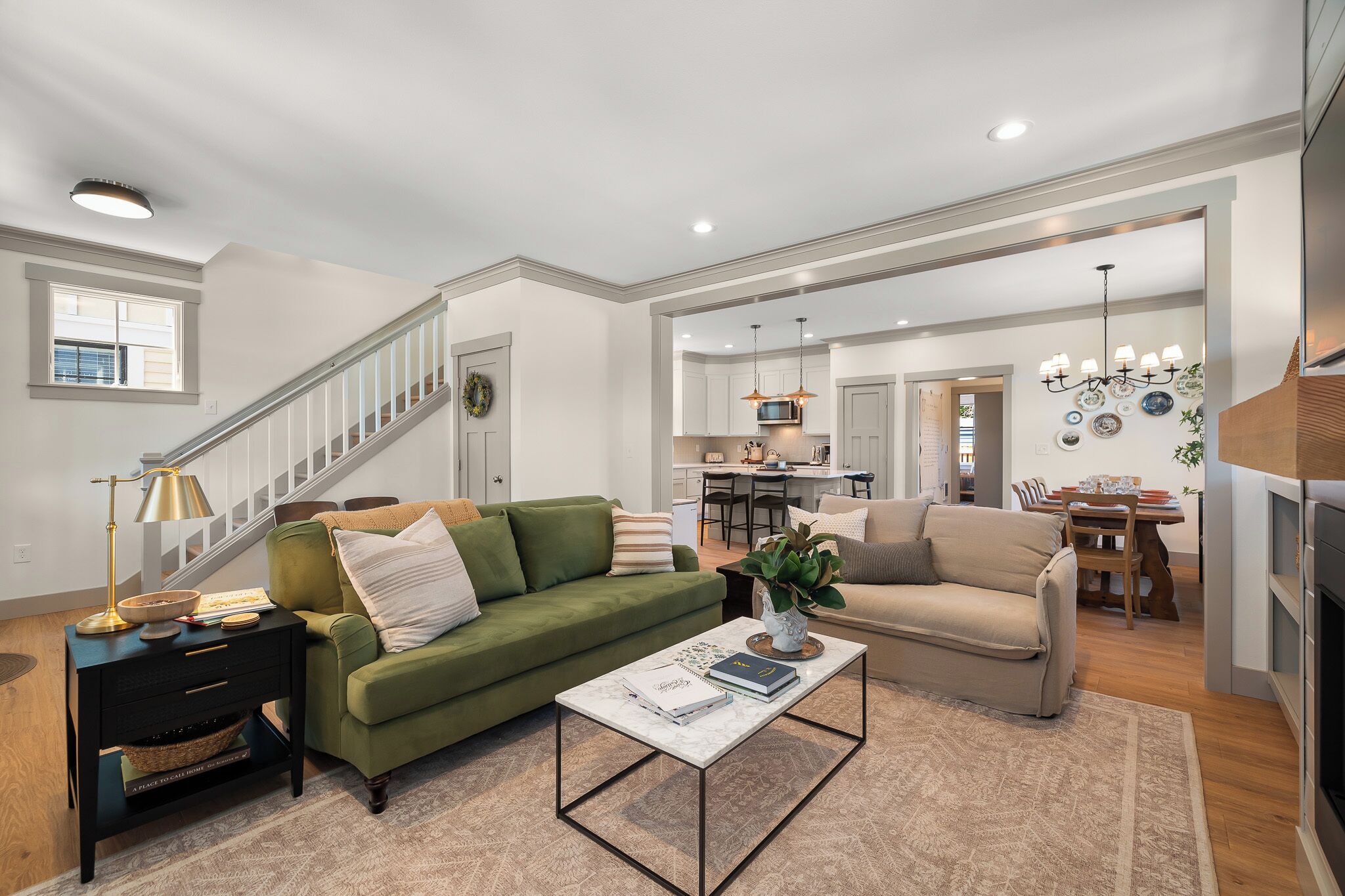 A spacious living room with a green sofa, a beige sofa, and a coffee table. There is a staircase leading to another level, a kitchen in the background, and a dining area.