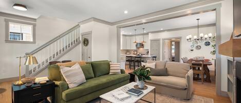 A spacious living room with a green sofa, a beige sofa, and a coffee table. There is a staircase leading to another level, a kitchen in the background, and a dining area.
