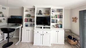 Un soggiorno moderno con una TV a schermo piatto, mobili bianchi, una ventola a soffitto e una piccola scrivania con una sedia.