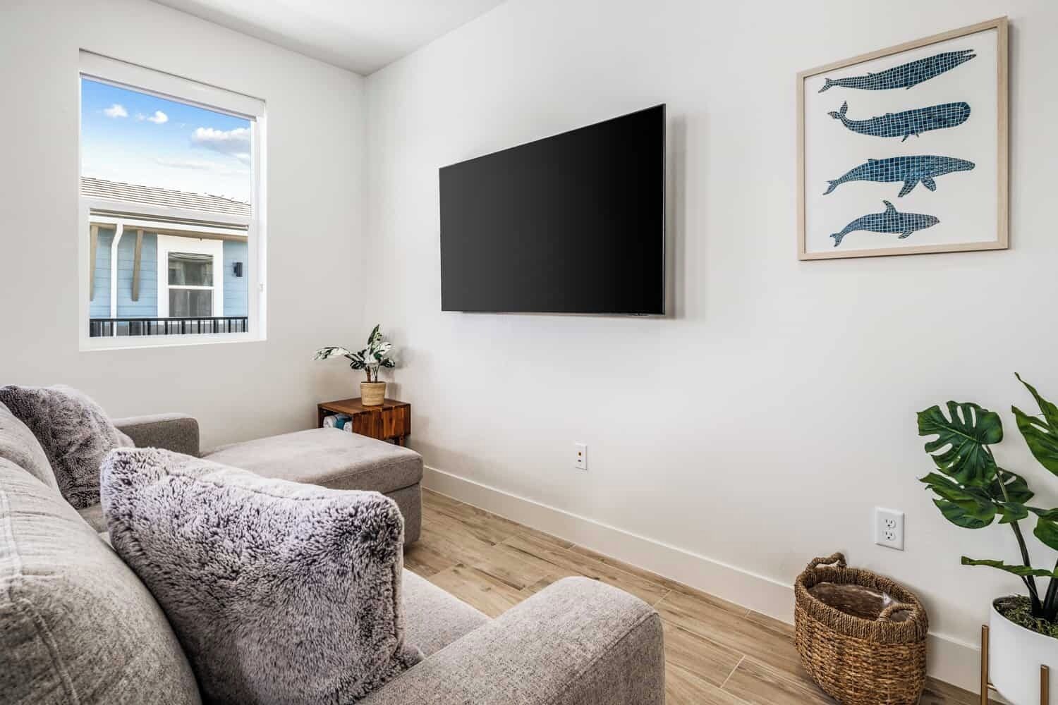 Una sala de estar moderna con un sofá gris, un televisor de pantalla plana montado en la pared y un ventanal con vistas a un edificio.