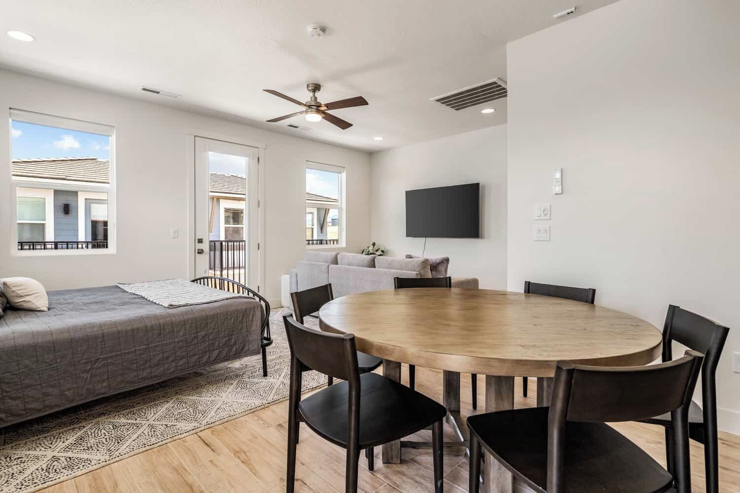 A modern room with a bed, a dining table with chairs, a ceiling fan, and a TV mounted on the wall.