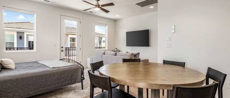 A modern room with a bed, a dining table with chairs, a ceiling fan, and a TV mounted on the wall.