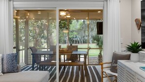 A room with a sliding glass door leading to a garden, a dining table with chairs, a sofa, and a small table with a plant. - Seabrook Exclusives | 1144 Summerwind (Johns Island)