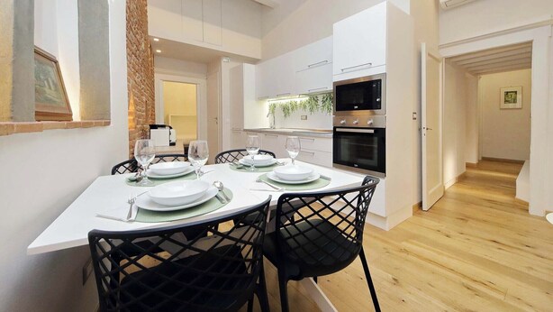 A modern kitchen with white cabinets, a built-in oven, and a dining area with a table set for four. - Santa Croce Gem w/ Chic Decor & Historic Charm (Florence)