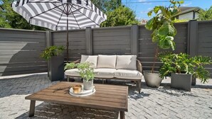 Un patio con una mesa de madera, un parasol a rayas, un sofá beige y plantas en macetas.