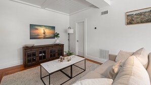 Una sala de estar con techo blanco, un mueble de madera, una mesa de centro blanca, un sofá beige y un cuadro de un atardecer sobre el océano.