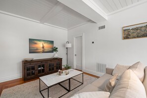 Una sala de estar con techo blanco, un mueble de madera, una mesa de centro blanca, un sofá beige y un cuadro de un atardecer sobre el océano.