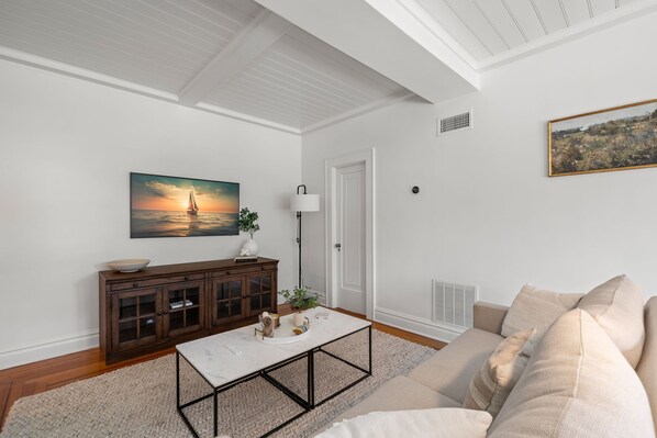 Una sala de estar con techo blanco, un mueble de madera, una mesa de centro blanca, un sofá beige y un cuadro de un atardecer sobre el océano.