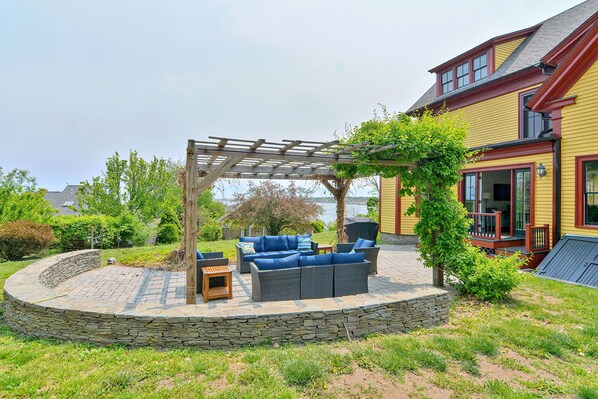 A covered outdoor seating area with stone walls and a pergola, surrounded by greenery and a view of a lake.