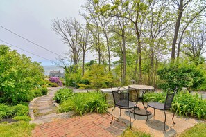 A garden patio with a round table and two chairs, surrounded by lush greenery and trees.
